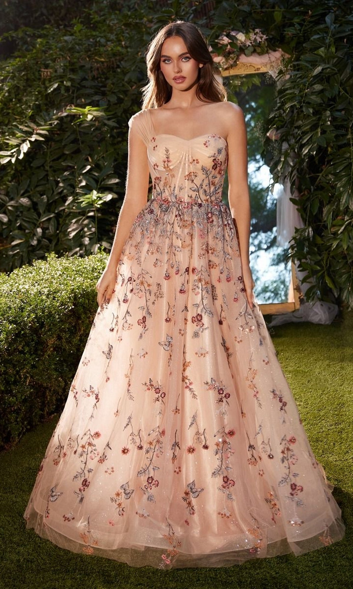 A woman stands on grass in a garden, wearing the Long Prom Dress A1289 by Andrea and Leo—a sleeveless, floor-length ball gown with thin straps, light pink hues, and floral embroidery. Green foliage and a mirror are in the background.
